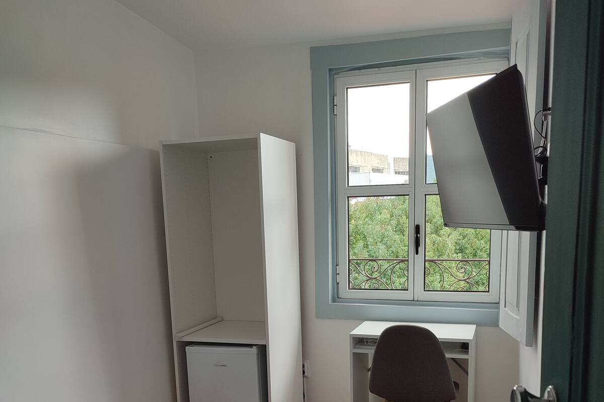 Een compacte kamer met een bed, een bureau en een stoel. Een raam biedt uitzicht op groen.