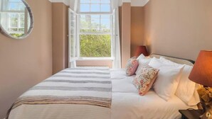 A bedroom with a bed, pillows, a bedside lamp, a round mirror, and a window with a view of greenery.