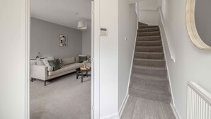 Un couloir avec un escalier menant à un deuxième étage, un mur de couleur gris clair, une porte blanche, un canapé gris avec des coussins, une petite table ronde et un luminaire fixé au mur.