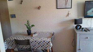 A small dining area with a table covered in a polka dot tablecloth, a potted plant, and a chair. There is a cabinet with a television and a lamp on top. - The DRYADES Mountain view (Gérardmer)