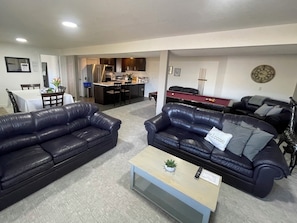 A living room with two black leather sofas, a coffee table, a pool table, and a kitchen area in the background.