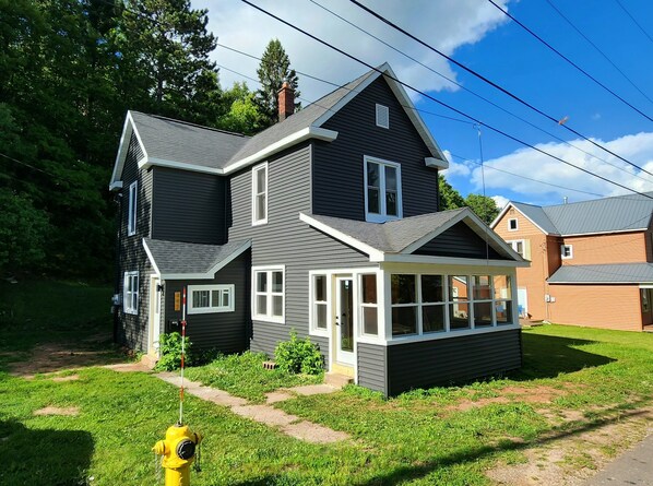 A two-story house with a dark exterior, white trim, and a covered porch. - Style-Ish 2 bed lower with all season sun room (Ishpeming)