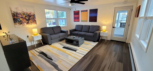 A modern living room with two grey sofas, a coffee table, and a patterned rug. There are two side tables with lamps, a ceiling fan, and a large abstract painting on the wall.