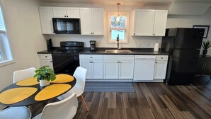 A modern kitchen with white cabinets, a black refrigerator, a microwave, and a stove. There is a dining area with a table and chairs, and a window with blinds.