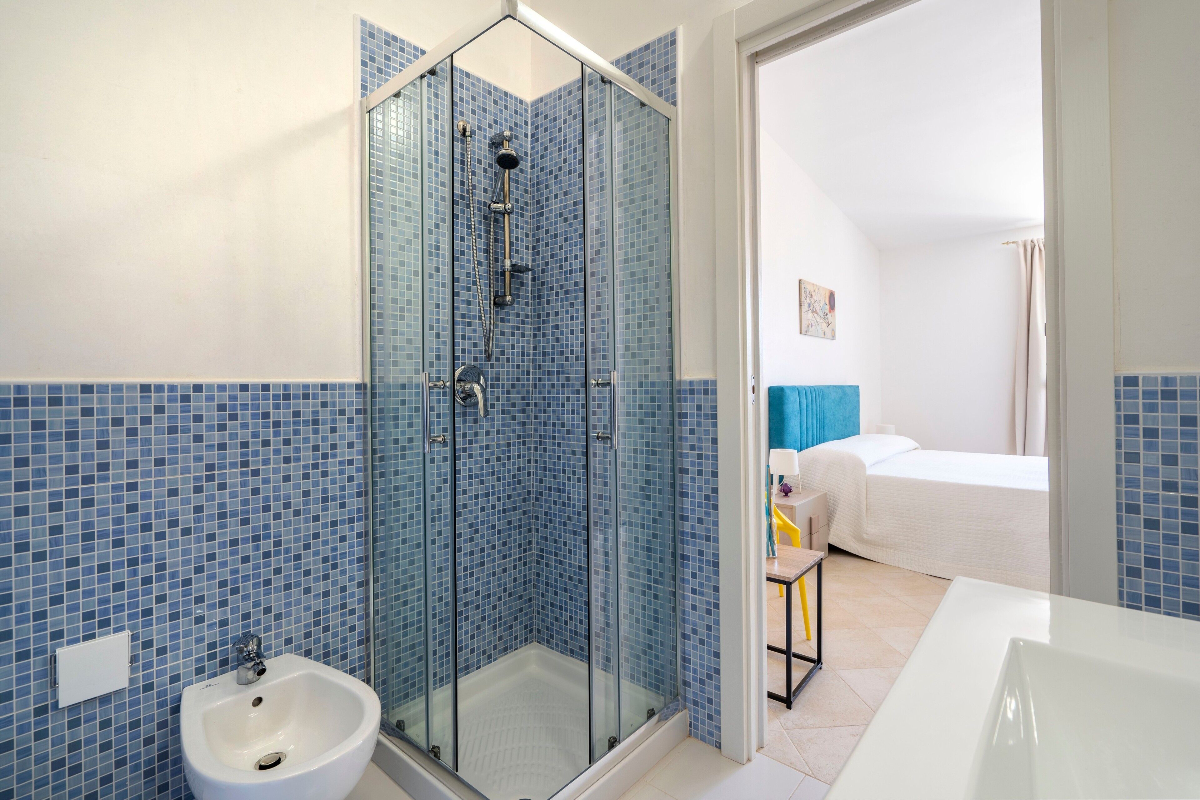 A bathroom with a glass-enclosed shower, a white sink, and a visible bed in the adjacent room.