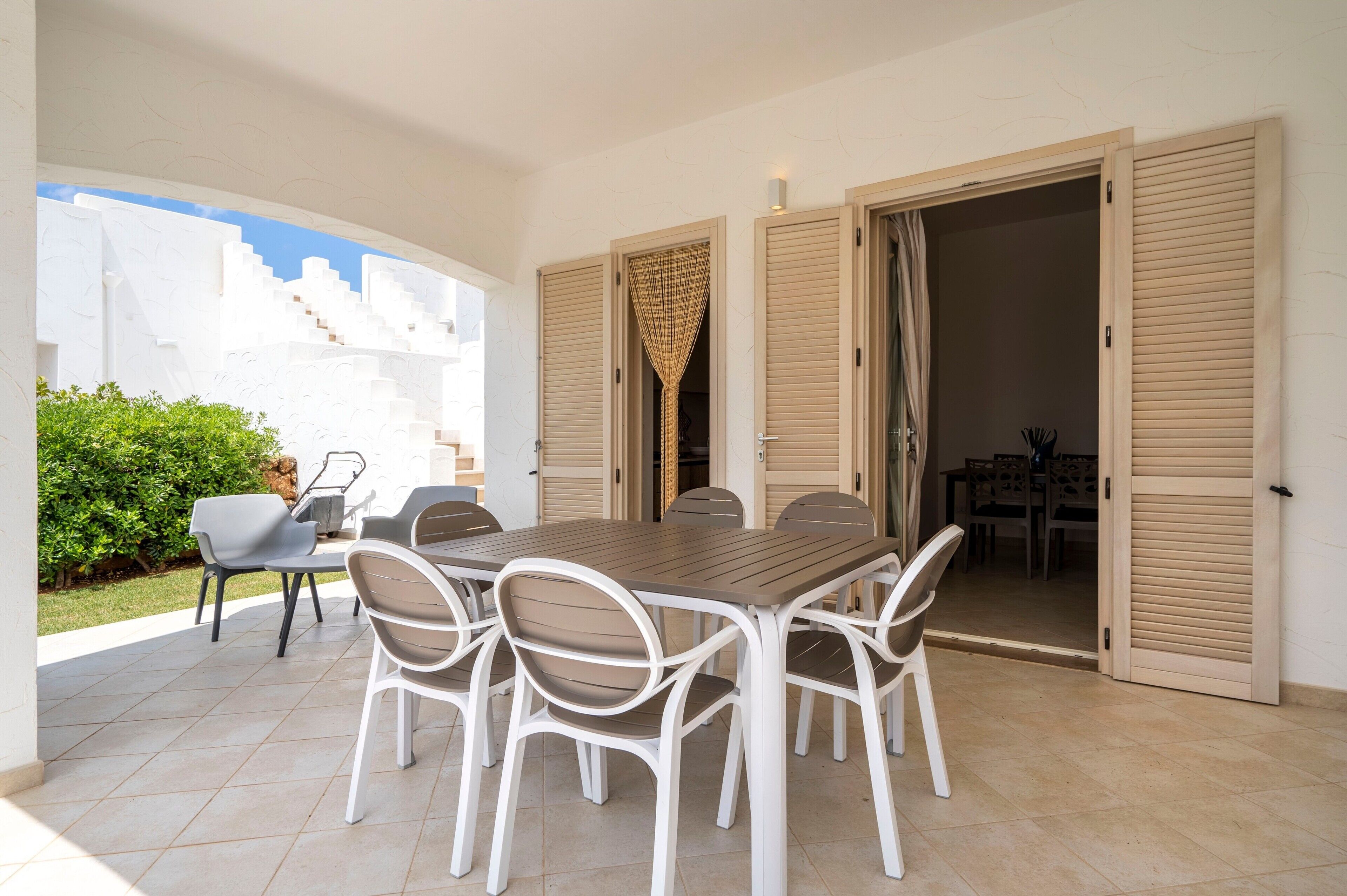 Eine Terrasse im Freien mit Esstisch und Stühlen, einer überdachten Sitzgelegenheit und Blick auf ein weißes Gebäude.