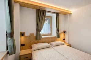 A bedroom with two beds, a window with curtains, a bedside table, and a wall-mounted light.