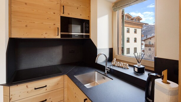 Una cucina con mobili in legno, un piano di lavoro nero, un lavello in acciaio inossidabile e una finestra con vista sugli edifici.