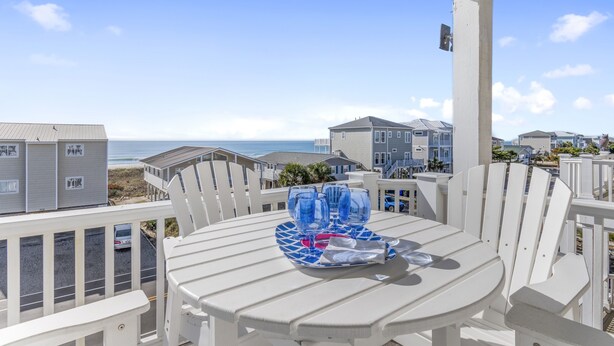 A balcony with a table set for two, overlooking a beach and a row of houses. - Makin' Memories! Clean, Comfortable, and Enjoyable! (Ocean Isle Beach)