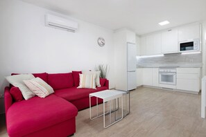 Un salon moderne avec un canapé rouge, une petite table blanche et un coin cuisine doté de meubles blancs et d’appareils électroménagers blancs.