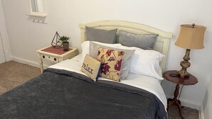Una cama bien hecha con una colcha gris oscuro, varios cojines incluyendo uno con la palabra 'relax' bordada, una mesita de noche con lámpara y un espejo fijado a la pared.
