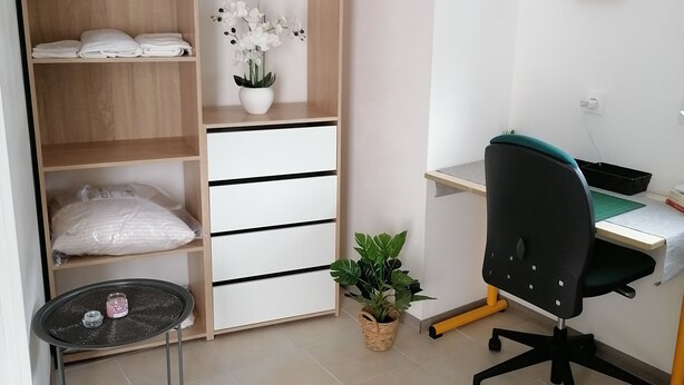 Quarto bem organizado com um guarda-roupa de madeira, uma mesa com cadeira, uma mesinha de centro com uma planta e uma prateleira na parede com objetos decorativos.