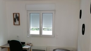 Quarto com cama, escrivaninha e cadeira. Há uma janela com persianas, um radiador e uma obra de arte pendurada na parede.