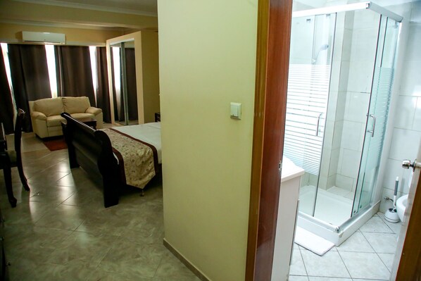 Habitación de hotel con cama, sofá, ducha con mampara de vidrio y baño con inodoro y lavamanos.