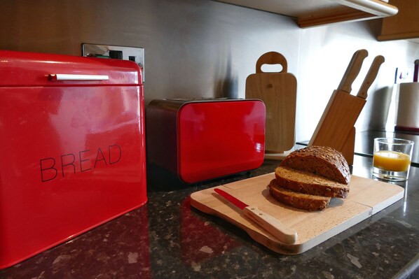 A kitchen counter with a red bread container, a toaster, a wooden cutting board with a loaf of bread, and a glass of orange juice. - Beachfront, Homely Apartment in Llandudno (Llandudno)
