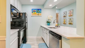 Una cocina moderna con armarios blancos, un refrigerador negro, un lavavajillas y un lavabo. En la pared, hay un cuadro de una playa y, sobre la encimera, un jarrón con flores.
