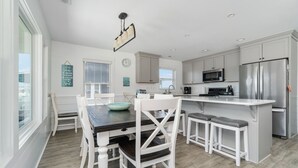 A modern kitchen with white cabinetry, a central island with stools, a stainless steel refrigerator, and a dining area with a dark wooden table and white chairs. - Impeccable home w/ pool on the desirable Ocean Ridge Drive of Atlantic Beach, NC (Atlantic Beach)