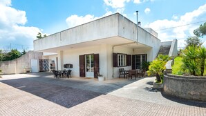 Un bâtiment moderne doté d’un toit plat, d’un patio couvert avec des sièges extérieurs et d’un grand bac à fleurs contenant des plantes tropicales.