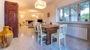 Un espace repas comprenant une table en bois, des chaises blanches, un luminaire suspendu et une fenêtre donnant sur un paysage verdoyant.