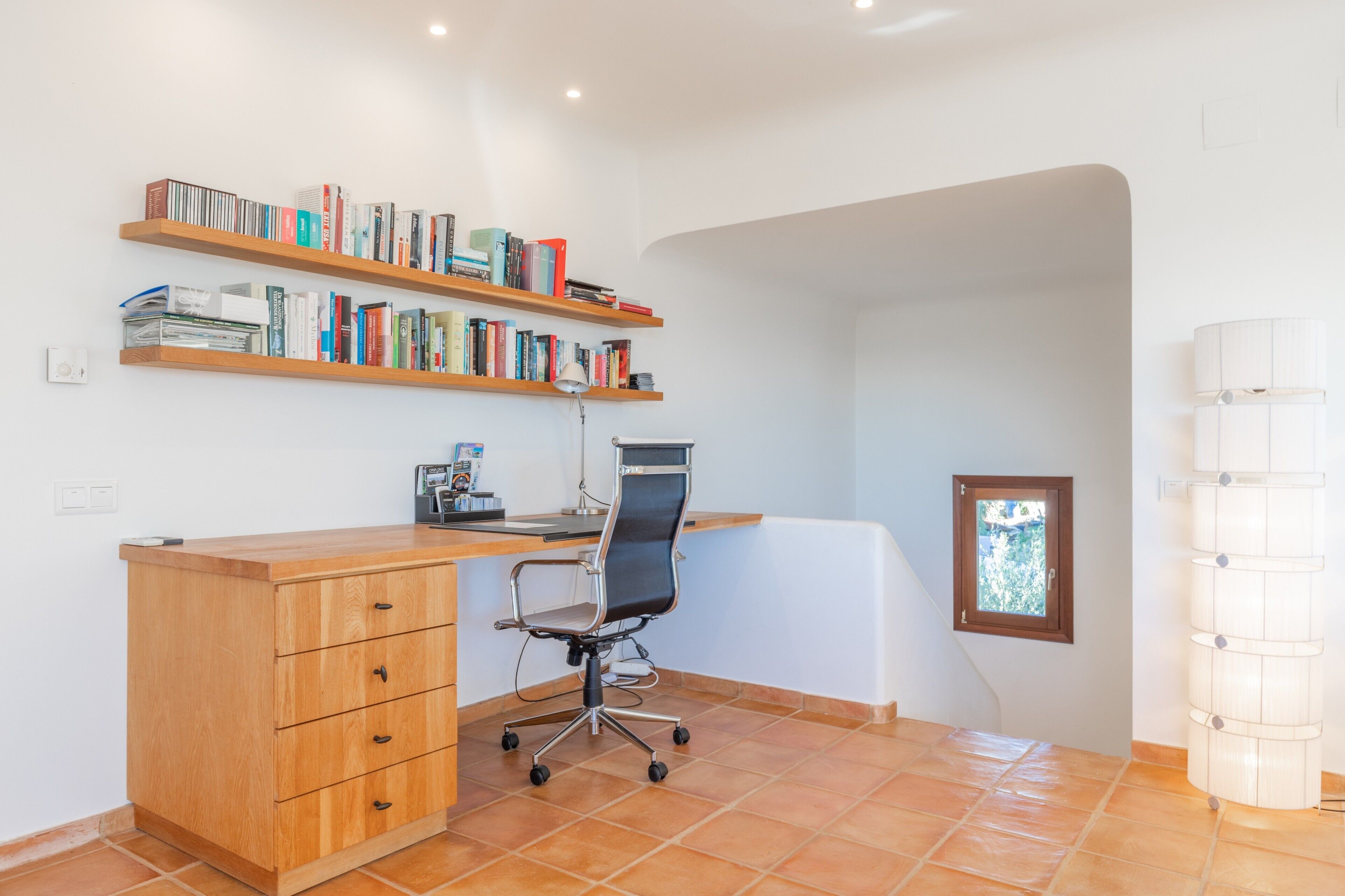 A room with a desk, chair, wooden shelves with books, a window, and a lamp.