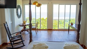 A bedroom with a large bed, a rocking chair, a table with a lamp, and a balcony with a view. - Domaine des tourterelles (Capesterre-de-Marie-Galante)