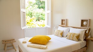 A neatly made bed with white bedding and a yellow pillow, a wooden nightstand with a lamp, and a window with open shutters. - Domaine des tourterelles (Capesterre-de-Marie-Galante)