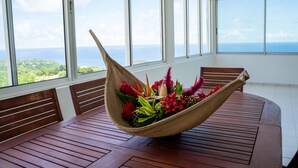 A wooden table with a woven basket filled with flowers, set in a room with large windows overlooking a scenic view. - Domaine des tourterelles (Capesterre-de-Marie-Galante)