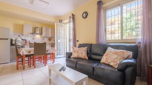Un salon comprenant un canapé, une table à manger, des chaises, une table basse, un coin cuisine et une grande fenêtre avec des rideaux.