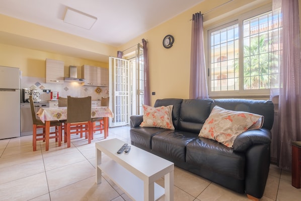 A living room with a sofa, a dining table, chairs, a coffee table, a kitchen area, and a large window with curtains.