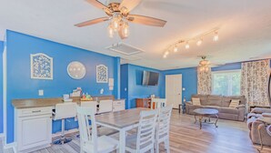 Una sala de estar luminosa con una pared azul, una mesa de comedor de madera, sillas blancas, un sofá y un ventilador de techo.