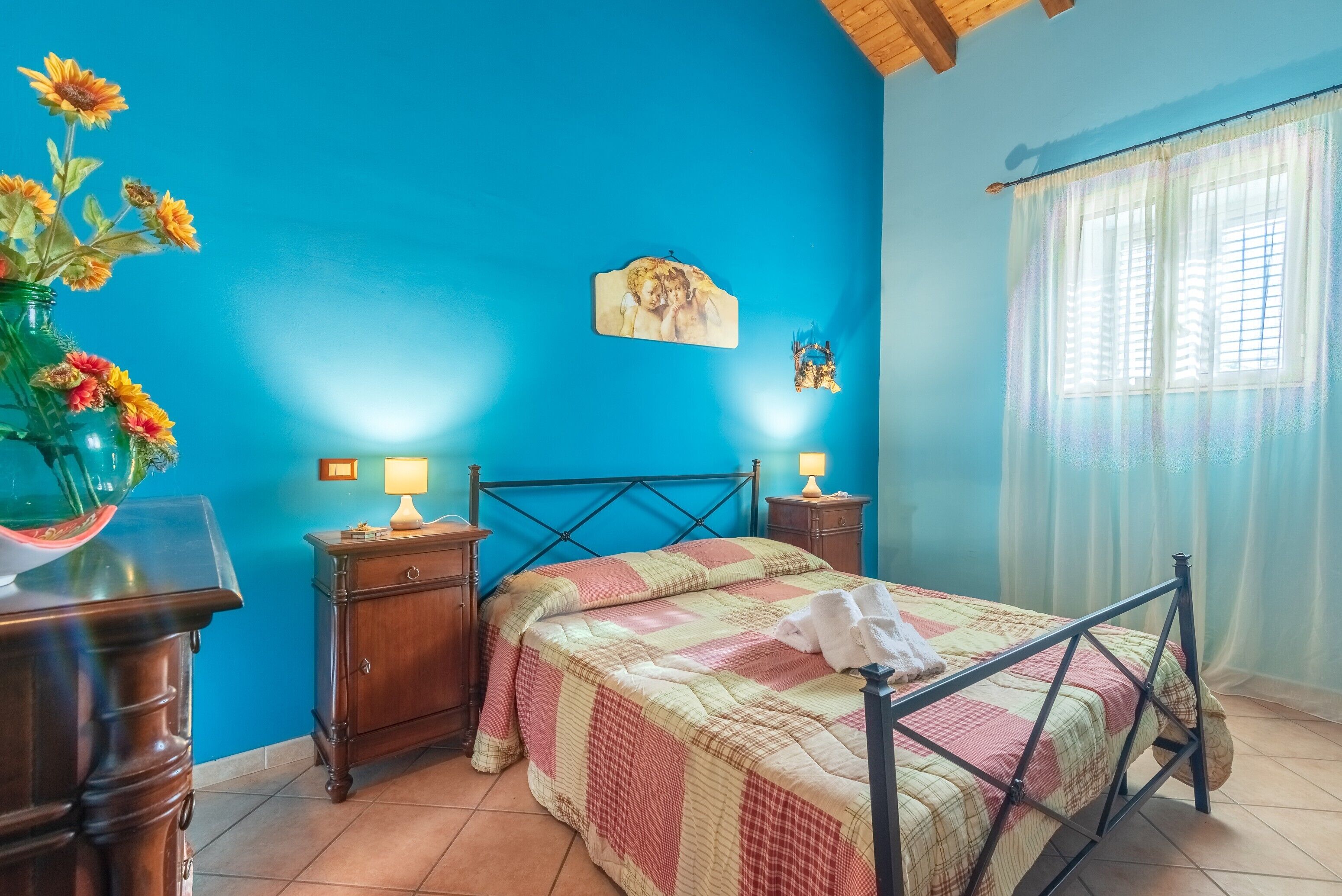 A bedroom with a bed, nightstands, a dresser, a vase of flowers, a wall clock, and a window with curtains.
