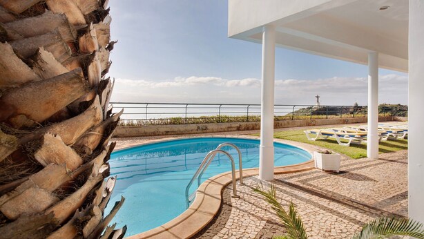 Ein Poolbereich mit freiem Blick auf das Meer, ein weißes Gebäude und eine Steinmauer.