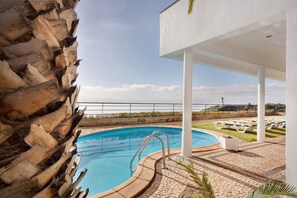 A pool area with a clear view of the sea, a white building, and a stone wall.