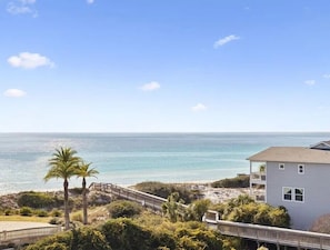 A coastal view with a beach, palm trees, and a house with a balcony overlooking the ocean.