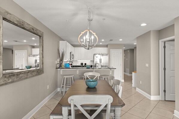 A dining area with a wooden table, white chairs, a chandelier, and a kitchen in the background.