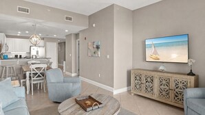 A modern living room with a round coffee table, grey sofas, a TV on the wall, and a kitchen in the background. - Gulf and Pool Views, Free Seasonal Beach Service, Designated Beach Area on 30A!! (Santa Rosa Beach)