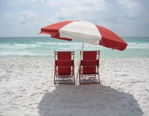 Two red beach chairs under a red and white beach umbrella on a sandy beach with the ocean in the background.