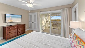 A bedroom with a bed, a ceiling fan, a dresser, a TV, and a view of a wooden deck and beach. - Gulf Front Townhome, Downtown Destin! (Destin)