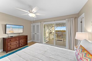 A bedroom with a bed, a ceiling fan, a dresser, a TV, and a view of a wooden deck and beach.
