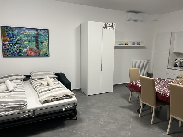 A modern bedroom with a bed, a wardrobe, a dining table with chairs, and a colorful abstract painting on the wall.
