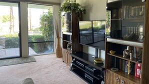Un salón con un televisor grande, estanterías de madera, un jarrón de mimbre y un ventanal corredero que da a un patio.