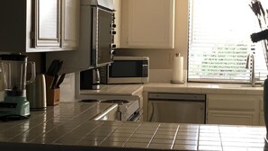 Una cocina con armarios blancos, un microondas, una licuadora y un ventanal con persianas.