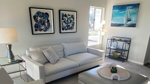 A modern living room with a white sofa, a round coffee table, and a grey armchair. There are two framed abstract paintings on the wall and a sailboat painting above a black metal shelf. - Coastal Condo w/Great Amenities, Walkable to Beach (Dana Point)