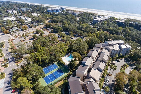 An aerial view of a residential area with a large swimming pool, tennis courts, and a beachfront.
