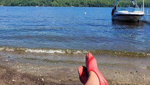 Vista del lago con un bote amarrado en el agua, una persona relajándose en la orilla y una colina boscosa al fondo.