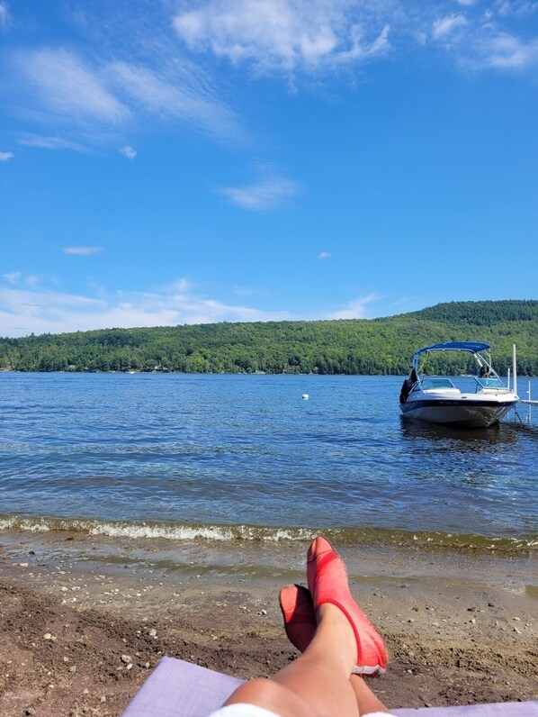 A lakeside view with a boat docked on the water, a person relaxing on the shore, and a forested hill in the background.