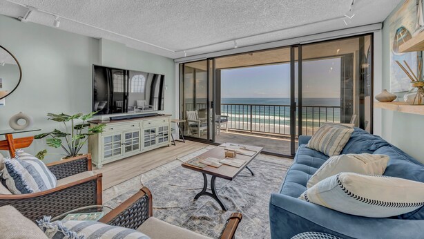 A spacious balcony with a view of the ocean, a comfortable seating area with a sofa and chairs, a flat-screen TV, and a cabinet with decorative items.