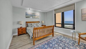 A bedroom with a wooden bed, nightstands, a small round table, a large window with a view of the ocean, and a patterned rug.