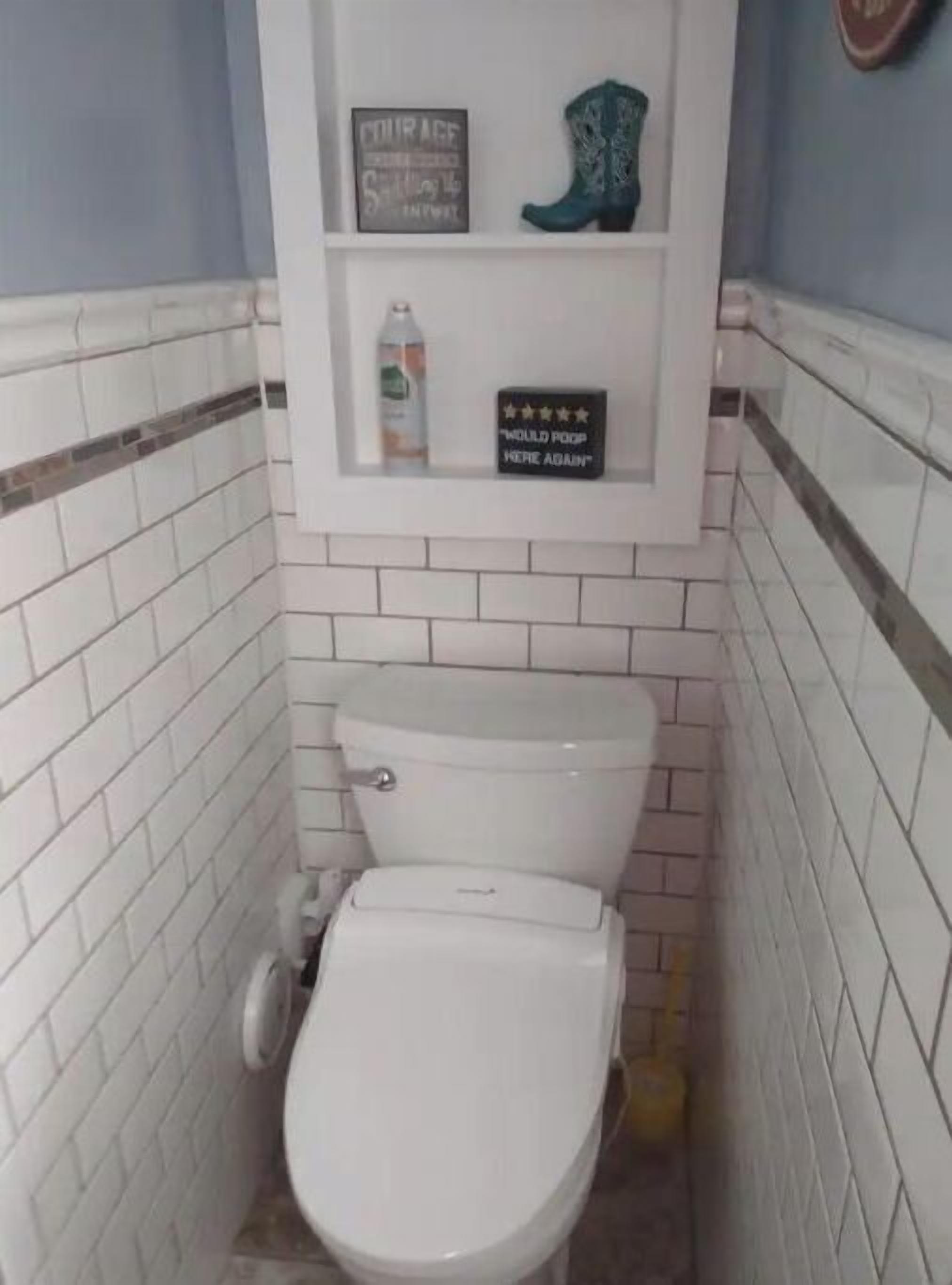 Un baño pequeño con paredes de azulejos blancos y un inodoro blanco. Hay un estante sobre el inodoro con un cartel que dice "Would poop here again" y un par de botas.