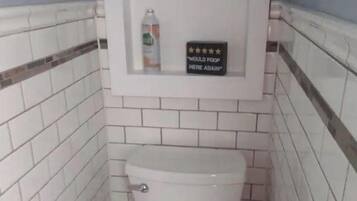 Un baño pequeño con paredes de azulejos blancos y un inodoro blanco. Hay un estante sobre el inodoro con un cartel que dice "Would poop here again" y un par de botas.